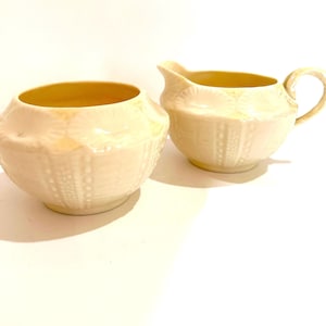 May include: A cream-colored ceramic sugar bowl and matching creamer pitcher set. Both pieces feature a textured, raised shell-like pattern on the exterior and a smooth, pale yellow interior finish. The creamer includes a curved handle and spout.