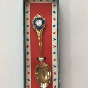 May include: A gold-colored souvenir spoon with a clock-shaped emblem, presented in a box with a red and white striped background and blue stars. The emblem reads "Bily Clocks Spillville, Iowa". Made in U.S.A.
