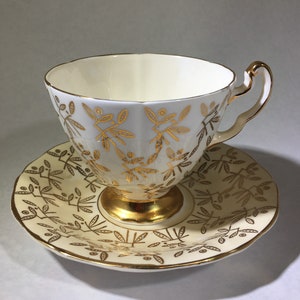 May include: A white teacup and saucer with a gold floral pattern. The cup has a fluted design and a gold rim. The saucer has a scalloped edge.