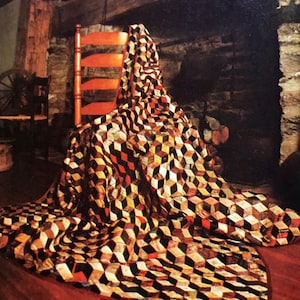 May include: A quilt with a geometric pattern of black, brown, gold, and red squares, draped over a wooden chair in front of a stone fireplace.