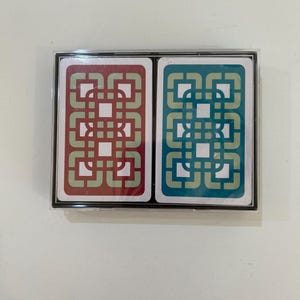 May include: A deck of playing cards in a clear plastic case. One card is red with a geometric design in a lighter shade of red. The other card is teal with a geometric design in a lighter shade of teal.