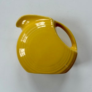May include: A yellow ceramic pitcher with a wide, rounded base and a single handle. The pitcher has a smooth, glossy finish and a subtle, rippled design.