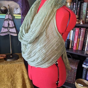 May include: A light green, crinkled scarf draped over a red dressmaker's form. The scarf is made of a lightweight fabric and has a flowing, textured appearance. The form is set against a backdrop of bookshelves and decorative items.