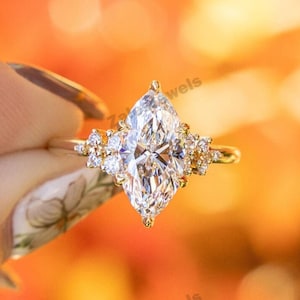 Könnte beinhalten: Ein goldener Verlobungsring mit einem großen, birnenförmigen Diamanten als Mittelpunkt, flankiert von kleineren Akzentdiamanten. Der Ring wird vor einem verschwommenen, warmfarbigen Hintergrund präsentiert, der das Funkeln der Edelsteine hervorhebt. Ein Symbol der Liebe.