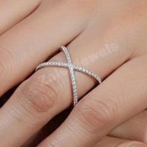 May include: A silver ring featuring a crisscross design, set with small, sparkling stones. The ring is worn on a finger, highlighting its delicate and elegant style. The ring's design offers a modern twist on a classic aesthetic.