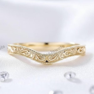 May include: A gold-coloured, curved wedding band featuring an elaborate, swirling filigree pattern. The ring is displayed on a white, silky surface, with several small, sparkling stones scattered around. The band has a V-shape.
