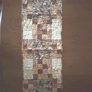 May include: A quilted table runner with a patchwork design in shades of brown, beige, and cream. The runner features a mix of geometric and floral patterns, with a rectangular shape and a textured appearance. The runner is approximately 122 cm long.