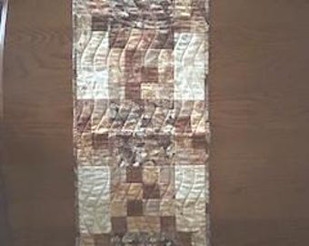 May include: A quilted table runner with a patchwork design in shades of brown, beige, and cream. The runner features a mix of geometric and floral patterns, with a rectangular shape and a textured appearance. The runner is approximately 122 cm long.