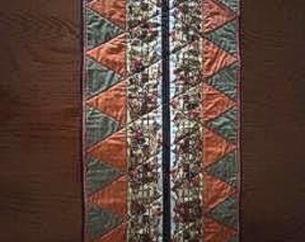 May include: A quilted table runner with a geometric design. The runner features alternating triangles in shades of orange, green, and a patterned fabric. A central strip of patterned fabric runs down the length of the runner.