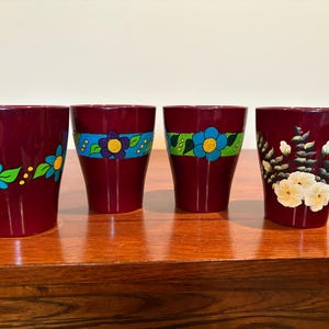 Könnte beinhalten: Vier weinrote Trinkgläser mit handgemalten Blumendesigns. Jedes Glas hat ein anderes Blumenmuster, darunter blaue und gelbe Blumen mit grünen Blättern. Die Gläser werden auf einer Holzoberfläche präsentiert.