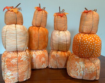 Può includere: Una collezione di zucche di stoffa in varie tonalità di arancione e bianco, disposte in formazioni impilate. Ogni zucca presenta un gambo e foglie decorative, perfette per l'arredamento autunnale. Le zucche sono esposte su una superficie di legno.