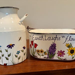 May include: Two white enamelware containers. One is a milk jug with handles and floral and bee designs. The other is a rectangular planter with the text "Earth Laughs in Flowers" and floral designs.