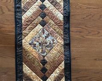 May include: A quilted table runner with a geometric design. The runner features a central diamond pattern surrounded by angled stripes in shades of brown, gold, and tan. A black border frames the design. The runner is placed on a wooden surface.