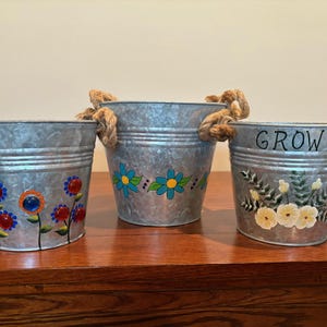 Puede incluir: Tres cubos de metal galvanizado con asas de cuerda. Los cubos están decorados con flores pintadas en varios colores. Un cubo tiene la palabra "GROW" pintada.
