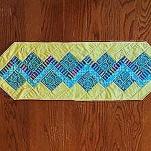 May include: A yellow quilted table runner with a diamond pattern. The diamonds alternate between blue and teal with patterned designs. The runner is approximately 12 inches wide and 48 inches long.