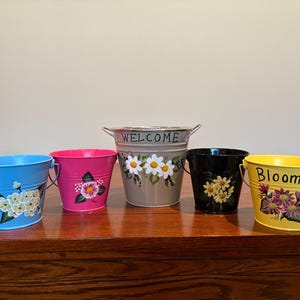 Może przedstawiać: Pięć kolorowych metalowych wiader z uchwytami, każde pomalowane we wzory kwiatowe. Wiadra są niebieskie, różowe, szare, czarne i żółte. Szare wiadro ma napis "WELCOME", a żółte wiadro ma napis "Bloom".