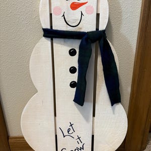 Puede incluir: Decoración de muñeco de nieve de madera con acabado pintado de blanco. Cuenta con una bufanda negra, detalles de botones negros y una nariz de zanahoria. El muñeco de nieve tiene mejillas rosadas y una boca sonriente. Las palabras "Let it Snow" están escritas en la parte inferior.