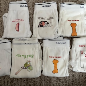 Pode incluir: Cuecas boxer brancas com vários desenhos gráficos e texto. Os desenhos incluem um cachorro-quente, uma personagem de desenho animado, um osso, o texto "Hot Stuff", um pônei, cartas de jogar e as frases "The Beast Within", "Bury the Bone", "Ride My Pony", "Bad to the Bone" e "Get Lucky".