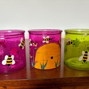 May include: Three glass candle holders in shades of pink and green, decorated with hand-painted bees and a beehive. The candle holders are cylindrical and sit on a wooden surface.