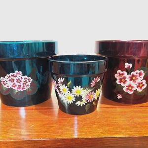 May include: Three black flower pots with hand-painted floral designs. The largest pot has pink and white flowers, the medium pot has white and pink daisies, and the smallest pot has pink and white flowers. The pots are on a wooden surface.