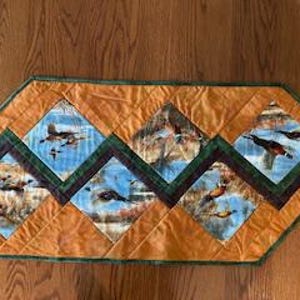 Puede incluir: Camino de mesa acolchado con un patrón de diamantes que presenta imágenes de patos en vuelo contra un cielo azul. El camino de mesa tiene un borde naranja y ribete verde, sobre una superficie de madera.