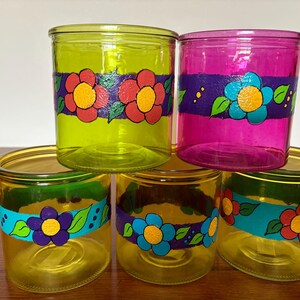 Pode incluir: Seis frascos de vidro empilhados em tons de amarelo, verde e rosa. Cada frasco apresenta um desenho floral pintado à mão em cores contrastantes. Os frascos estão colocados sobre uma superfície de madeira.