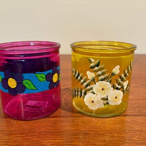May include: Two hand-painted glass votive candle holders. One is pink with a floral design in blue, purple, and green. The other is yellow with white flowers and green leaves. Decorative home decor.