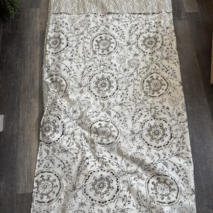 May include: A rectangular, folded textile featuring a floral pattern in shades of gray and black on a white background. The top section has a chevron design in light gray. The fabric is lying on a wooden surface.