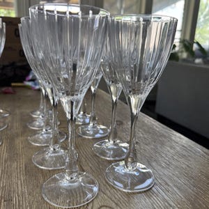 May include: A collection of clear, stemmed wine glasses with vertical ribbed detailing. The glasses have a classic shape with a wide bowl and a slender stem. The glasses are arranged on a wooden surface, suggesting a dining or entertaining setting.
