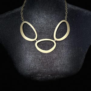 May include: A gold-coloured necklace with three linked oval pendants. The necklace is presented on a black mannequin, highlighting its design. The chain is fine, and the overall style is modern and minimalist, suitable for various outfits.