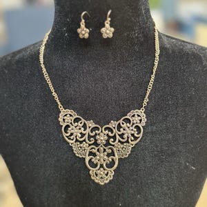 May include: A gold-colored necklace and earring set. The necklace features an ornate, filigree pendant with floral and scroll designs. The matching earrings are small flower-shaped studs. The jewelry is displayed on a black mannequin.