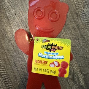 Op de afbeelding: Een rode Redberry marshmallow snoep van Sour Patch Kids in de vorm van het merkmascotte. De snoep weegt 54g en heeft een lachend gezicht. Een geel label is aan de snoep bevestigd.