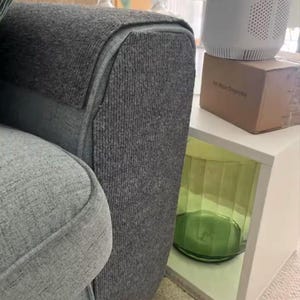 May include: Close-up of a gray upholstered sofa with a textured fabric. A white side table holds a white appliance and a cardboard box. A green glass vase is visible in the table's open compartment.