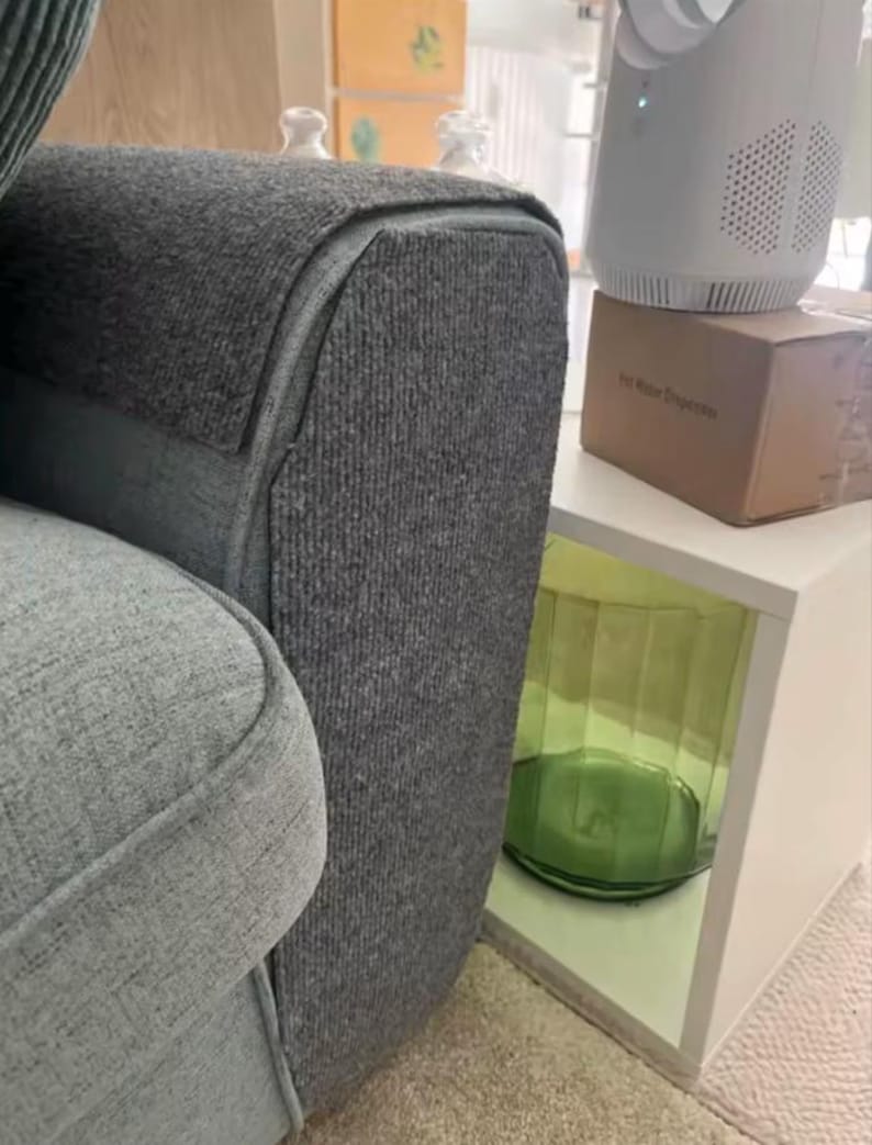 May include: Close-up of a gray upholstered sofa with a textured fabric. A white side table holds a white appliance and a cardboard box. A green glass vase is visible in the table's open compartment.