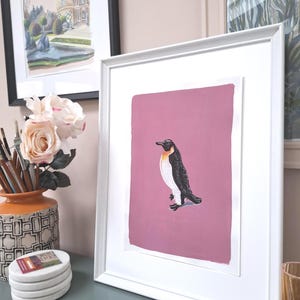 May include: Framed art print of a penguin on a pink background. The penguin is black and white with an orange chest. The print is in a white frame, and other framed art and decor are visible in the background.