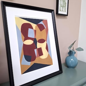 May include: Abstract art print in a black frame. The artwork features geometric shapes in shades of red, yellow, and blue. A white mat surrounds the art. A small blue vase with greenery sits nearby on a teal surface.