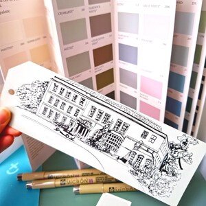 May include: A black and white architectural illustration of a building on a white tag, held in front of paint colour swatches. Three beige Micron 05 pens and a small paint swatch are also visible. The paint swatches include colours like Cromarty and Great White.