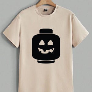 May include: Beige t-shirt featuring a black graphic of a jack-o'-lantern-style Lego head. The design has a smiling face with triangular eyes and a jagged mouth. The shirt has short sleeves and a crew neck, perfect for Halloween.