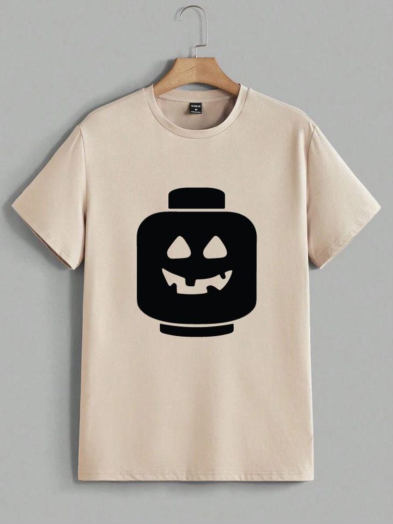 May include: Beige t-shirt featuring a black graphic of a jack-o'-lantern-style Lego head. The design has a smiling face with triangular eyes and a jagged mouth. The shirt has short sleeves and a crew neck, perfect for Halloween.