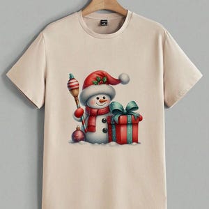 Pu&ograve; includere: T-shirt beige con un'illustrazione ad acquerello di un pupazzo di neve con cappello e sciarpa rossi di Babbo Natale. Il pupazzo di neve tiene una scopa a strisce e si trova accanto a un regalo confezionato con un fiocco verde acqua. &Egrave; presente anche un ornamento natalizio.