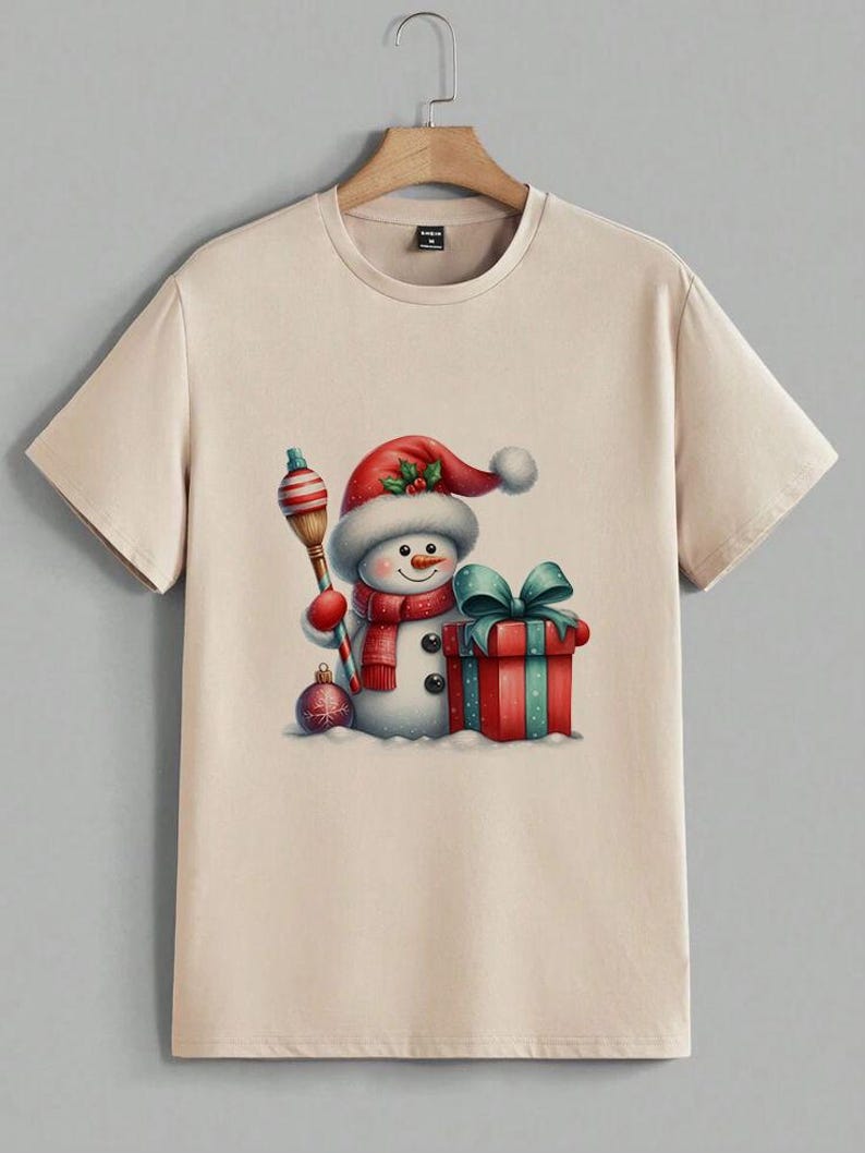 Pu&ograve; includere: T-shirt beige con un'illustrazione ad acquerello di un pupazzo di neve con cappello e sciarpa rossi di Babbo Natale. Il pupazzo di neve tiene una scopa a strisce e si trova accanto a un regalo confezionato con un fiocco verde acqua. &Egrave; presente anche un ornamento natalizio.