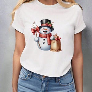 Pu&ograve; includere: T-shirt bianco con un allegro design a pupazzo di neve. Il pupazzo di neve indossa un cappello a cilindro nero, una sciarpa rossa e tiene in mano un rametto con un fiocco rosso. Un sacchetto di carta marrone con regali completa il look festivo.