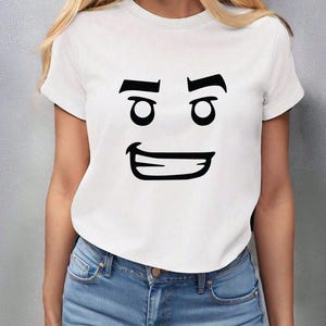 May include: White t-shirt with a black cartoon face graphic. The graphic features two eyes, eyebrows, and a smiling mouth. The shirt is paired with blue jeans.