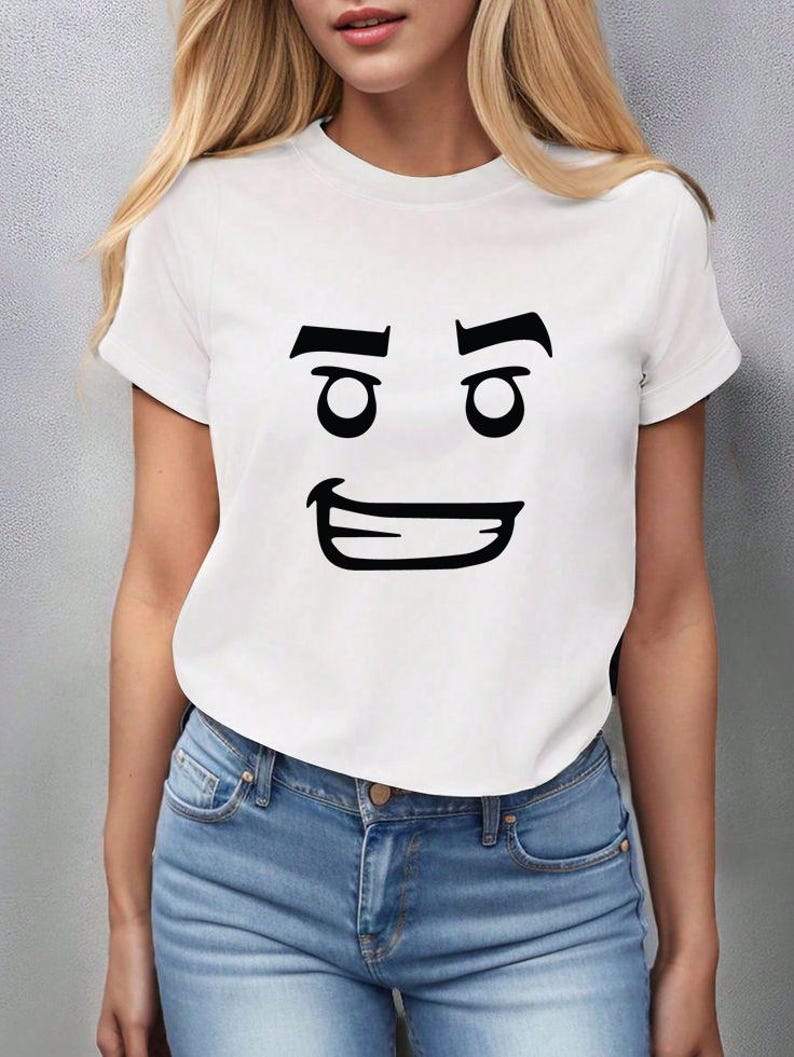 May include: White t-shirt with a black cartoon face graphic. The graphic features two eyes, eyebrows, and a smiling mouth. The shirt is paired with blue jeans.