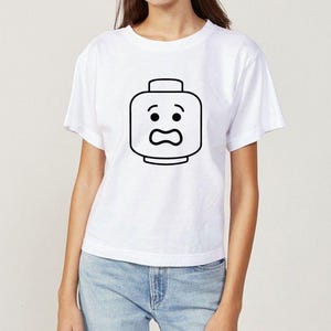 May include: White t-shirt featuring a black outline of a Lego head with a surprised expression. The graphic is centered on the front of the shirt. The shirt has short sleeves and a crew neck.