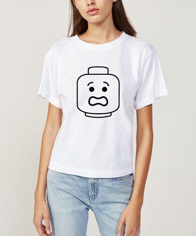 May include: White t-shirt featuring a black outline of a Lego head with a surprised expression. The graphic is centered on the front of the shirt. The shirt has short sleeves and a crew neck.