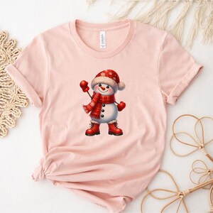 Pu&ograve; includere: T-shirt rosa chiaro con una grafica di pupazzo di neve allegro. Il pupazzo di neve indossa un cappello, una sciarpa, guanti e stivali rossi. La grafica &egrave; centrata sulla maglietta, creando un design festoso. La maglietta &egrave; in materiale morbido.