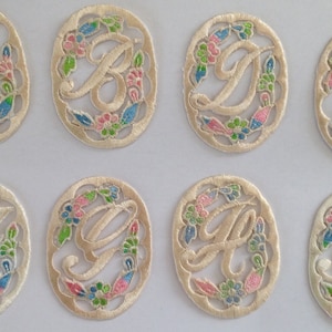 May include: Eight embroidered patches featuring floral designs and letters A through H in a cursive font. The patches are cream coloured with blue, green, and pink floral embroidery.