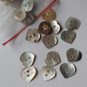 Puede incluir: Una colección de 14 botones de nácar en forma de corazón con dos agujeros cada uno. Los botones son de color gris claro iridiscente.