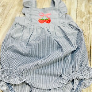 May include: Blue and white striped baby romper with a cherry applique. The romper features ruffled straps and leg openings. The cherry design has red cherries and a pink bow. The fabric has a seersucker texture.